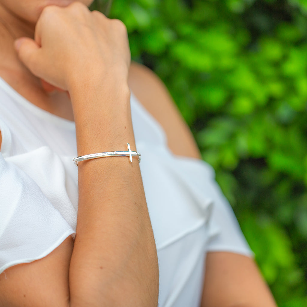Young woman wearing our Christian bracelet called Double-Edged Sword in sterling silver in the format of a sword and a Cross with "The Watchfulness of God" engraved on it. She is wearing a white blouse, standing in front of green bushes and has her hand on her chin.