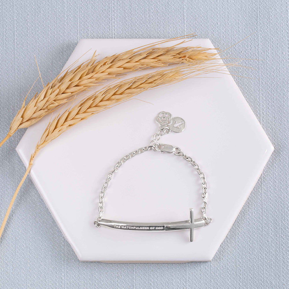 A Christian bracelet called Double-Edged Sword in sterling silver in the format of a sword and a Cross with "The Watchfulness of God" engraved on it, displayed on a white tile on a light blue background with two stems of wheat.