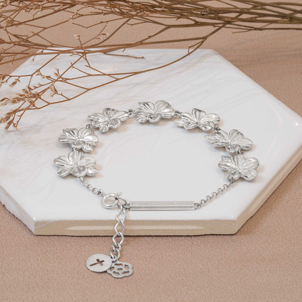 A Christian bracelet crafted from sterling silver, carrying a biblical significance , featuring seven exquisite almond flower buds and blossoms as a representation of God's watchful care over our lives on a white tile with dried flowers and light brown fabric.
