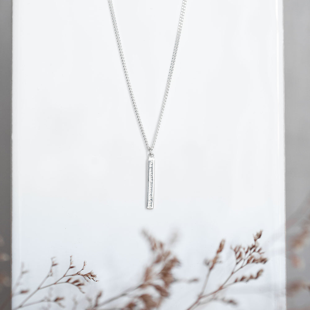 The Watchfulness of God engraved on a sterling silver bar necklace  serves as a reminder of God's watchful and caring presence in your life. Christian necklace with and extension of the cross of Jesus and our logo of an almond blossom on a white tile background with dried flowers