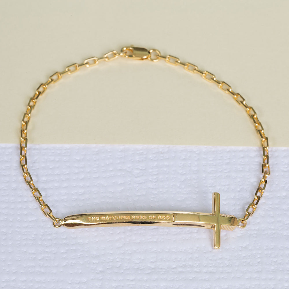 Christian bracelet in yellow gold in the format of a sword and a cross with "The Watchfulness of God" written on it. The bracelet is on a piece write textured paper and a green one
