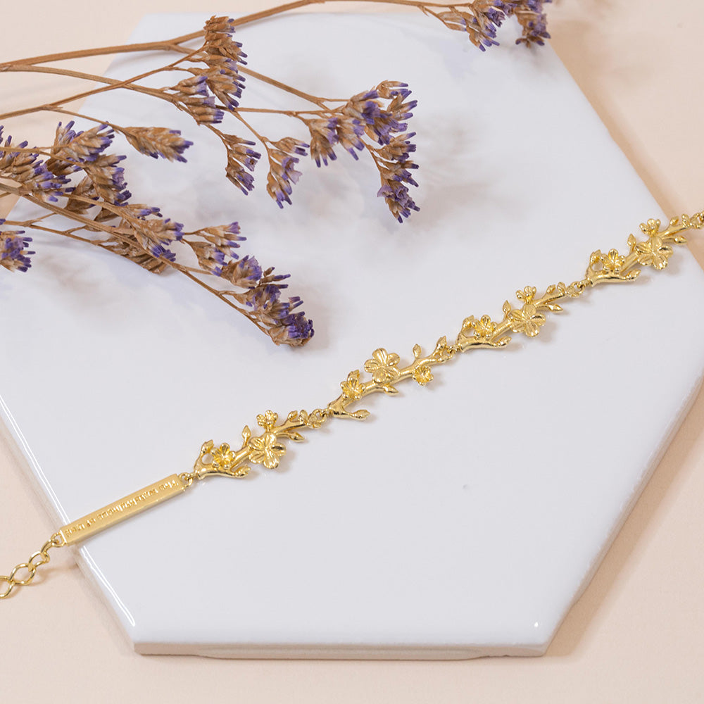 Our Esther 22 small almond flowers buds and  blossoms Christian bracelet in yellow gold plated over sterling silver displayed on a white tile with purple flower accents.