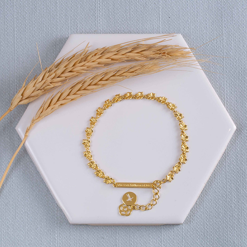 Our Hannah 22 almond blossoms Christian bracelet in yellow gold plated with our watchfulness of God bar, extension, logo and the cross displayed on a white tile with two stems of wheat on a light blue fabric background