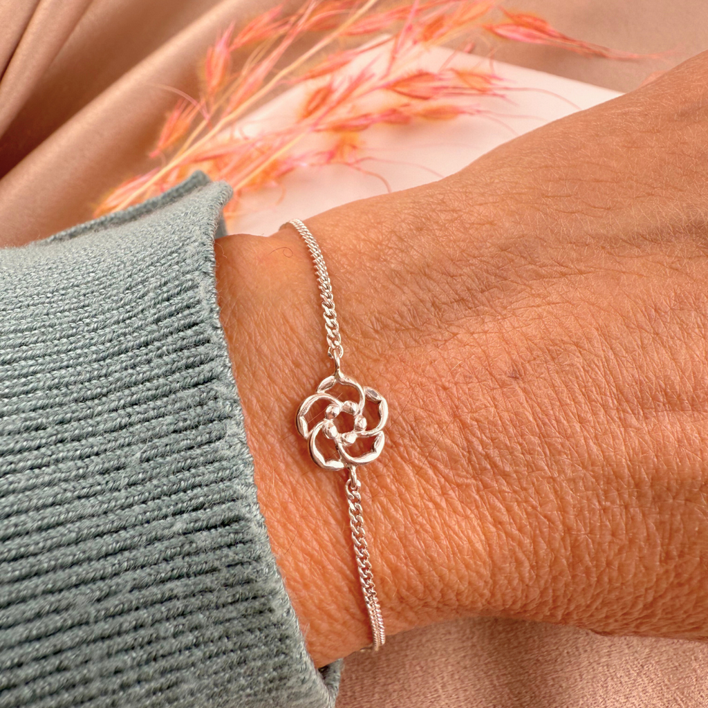 Silver bracelet with a floral charm on a wrist, with a blurred background of flowers and fabric.