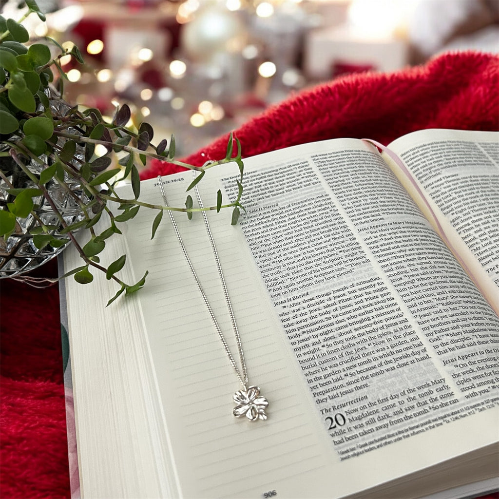 Open book with a silver necklace on a red surface, surrounded by greenery and blurred lights.