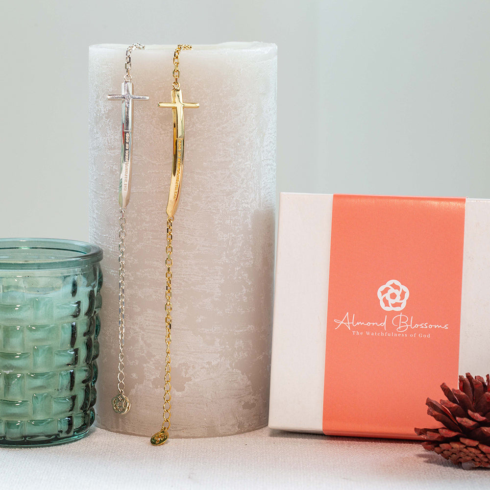 Two Christian bracelets called Double-Edged Sword  in sterling silver and gold plated in the format of a sword and a Cross with "The Watchfulness of God" engraved on it, displayed on a grey candle next to our brand packaging and a textured green candle.