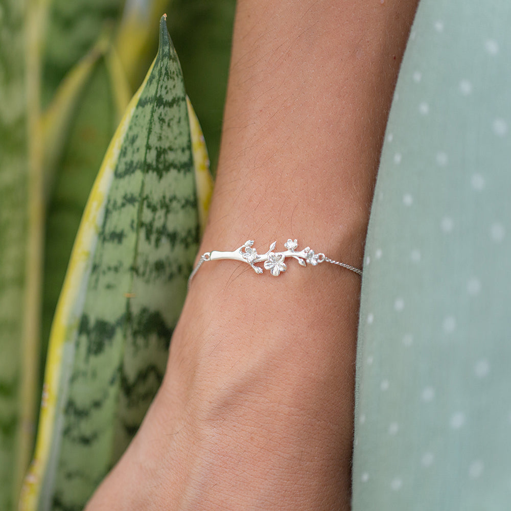 Young woman wearing our christian bracelet Esther, The Branch of an Almond Tree, in sterling silver with a single branch of an Almond Tree with its buds and blossoms. She is wearing a green dress with little white dots and there are plants on her left side on the background.