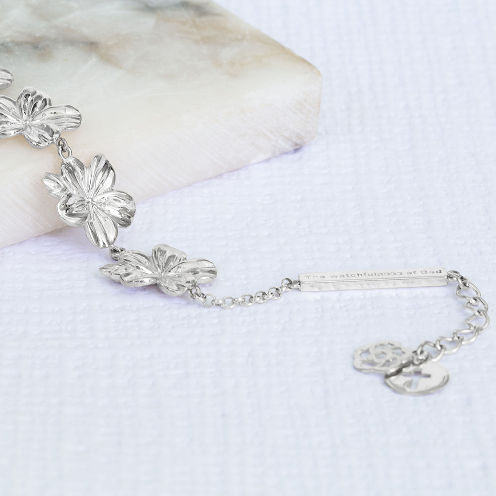 Mary Magdalene 7 christian bracelet in sterling silver, inspired by the lampstand of the tabernacle with 7 large almond blossoms and buds, 7 being the biblical number of completion and perfection, a bar with "The Watchfulness of God" written on it and two pendents, one with the cross and the other with our logo. The bracelet is on a piece of marble and a write textured paper.