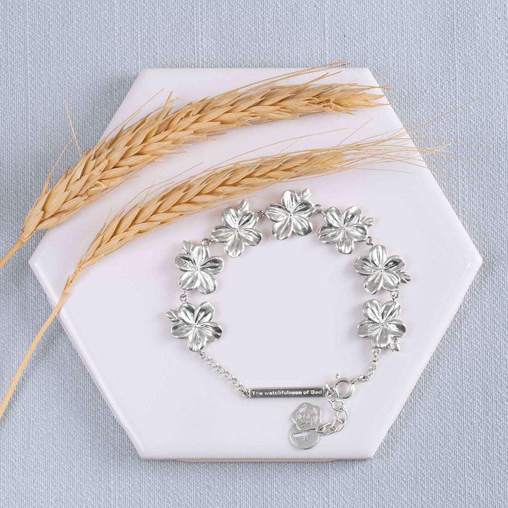 A Christian bracelet crafted from sterling silver, carrying a biblical significance , featuring seven exquisite almond flower buds and blossoms as a representation of God's watchful care over our lives on a white tile with wheat on a pale blue fabric