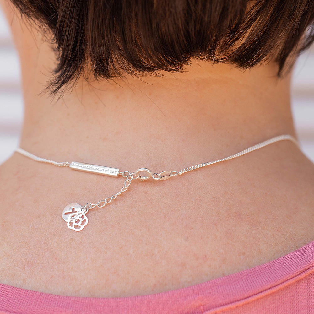 Our Esther Christian necklace the branch of an almond tree in sterling silver showcasing our watchfulness of God bar, extension with logo and the cross dangling off the back of the neck of a short dark haired woman wearing a pink shirt