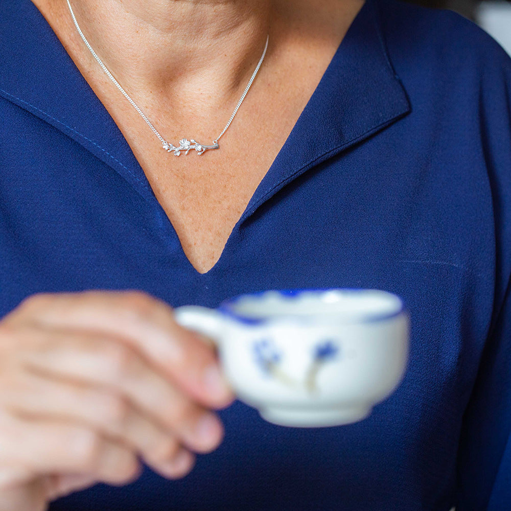  Our Esther Christian necklace, inspired by the branch of an almond tree,  in sterling silver on the neck of a woman holding a blue and white coffee cup wearing a navy blue blouse