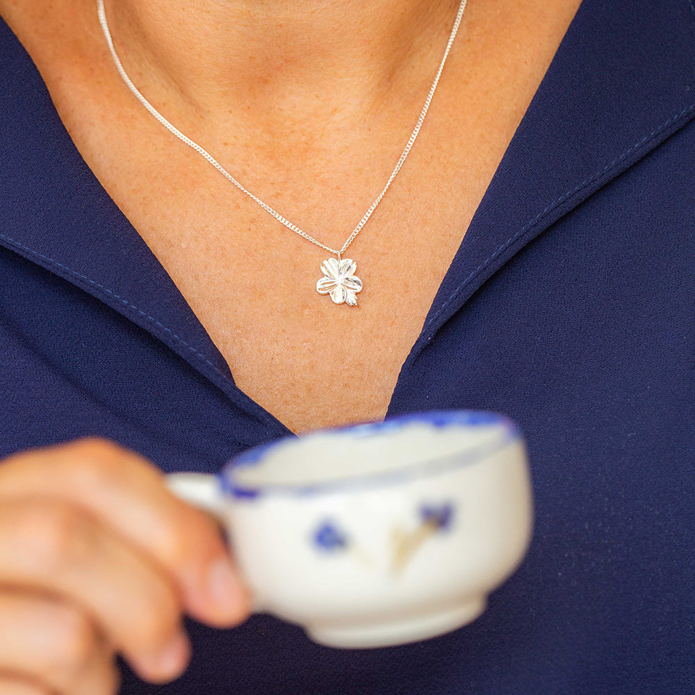 Our Mary Magdalene Christian necklace of an almond blossom and bud in sterling silver inspired by the meaning of the almond blossom in the Word of God and serves as a reminder of His watchfulness over your life. Pictured on a woman in a navy blue blouse holding a cup of coffee.