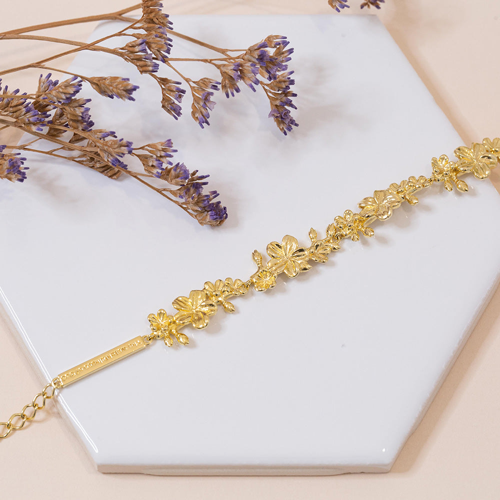 Our Esther 22 large almond blossoms Christian bracelet in yellow gold plated over sterling silver displayed on a white tile with purple flower accents.