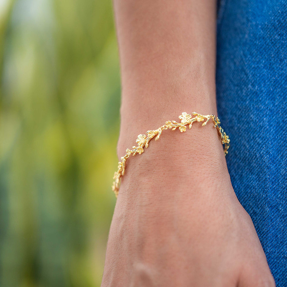 Young woman wearing our christian bracelet Esther 22S in yellow gold in the shape of branches with 22 almond buds and blossoms. She is wearing a jeans skirt and there are green plants out of focus background. 