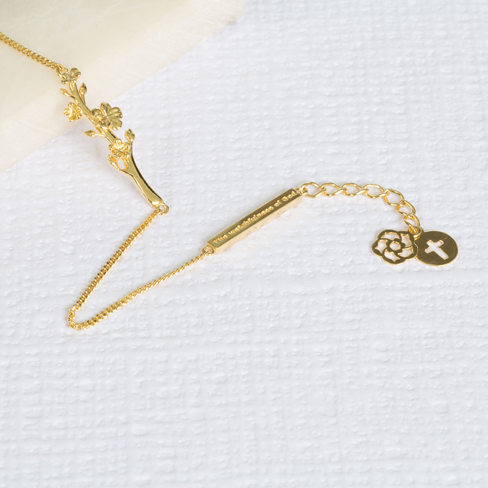 Esther, The Branch of an Almond Tree, christian bracelet in yellow gold with a single branch of an Almond Tree with its buds and blossoms, a bar with "The Watchfulness of God" written on it and two pendents, one with the cross and the other with our logo. The bracelet is on a piece of marble and a write textured paper.