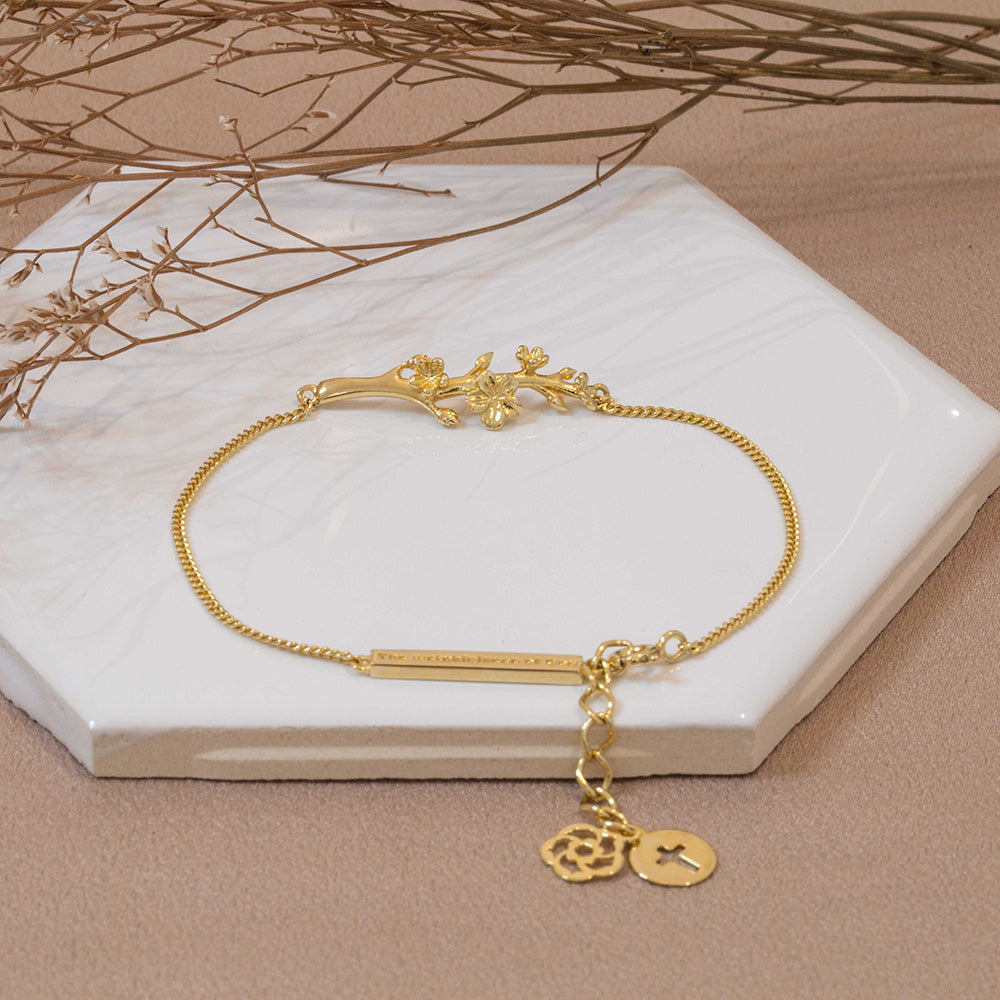 Our Esther  branch of an almond tree Christian bracelet gold plated over  sterling silver, with our extention of the Cross of Jesus and our logo of an almond blossom displayed on a white tile with tiny branches on a brown fabric background