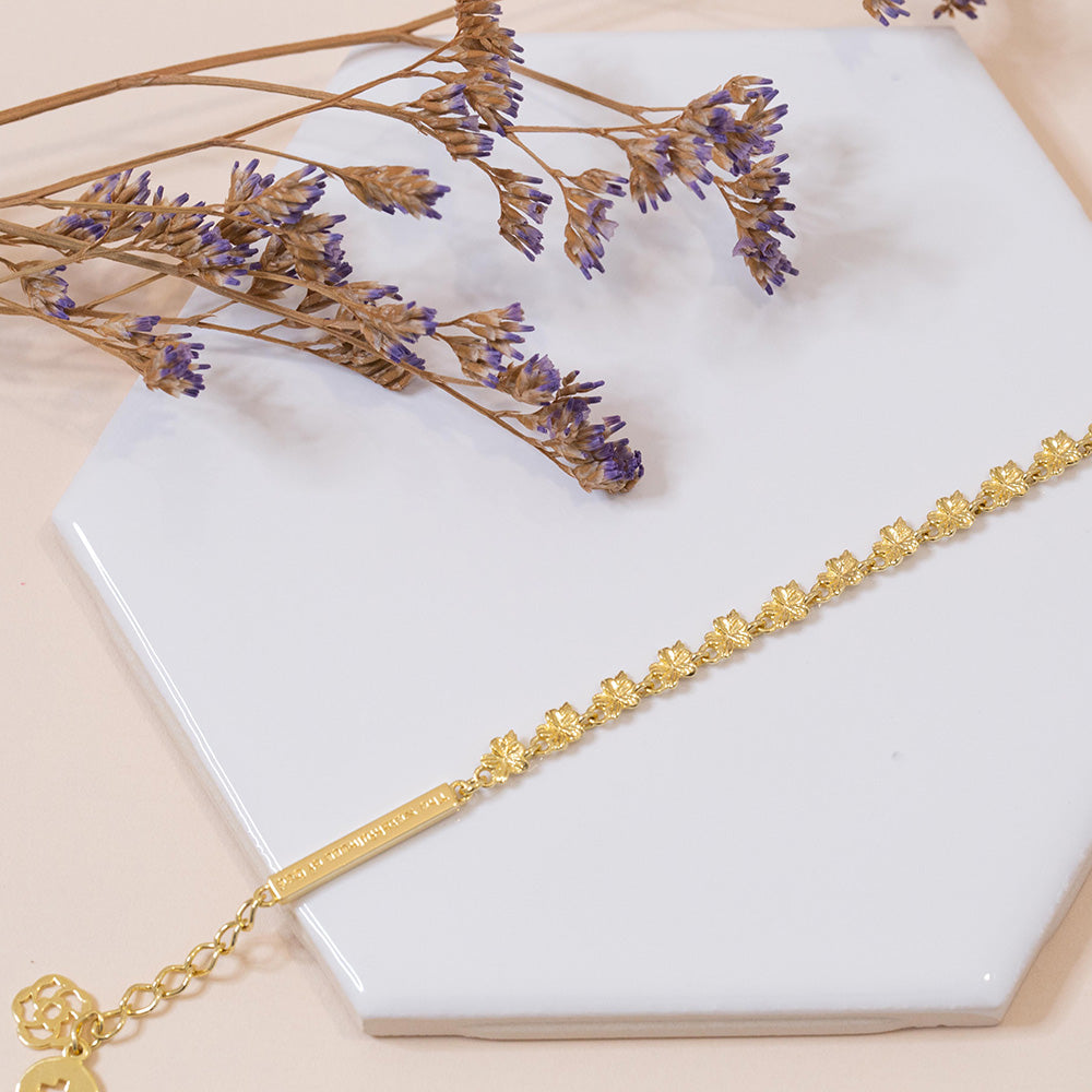 Our Hannah 22 almond blossoms Christian bracelet in yellow gold plated with our watchfulness of God bar, extension, logo, and the cross displayed on a white tile with small dried purple flower stems  on a pale pink fabric background.