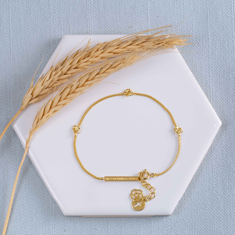 Hannah 3 christian bracelet in yellow gold plated, inspired by the lampstand of the tabernacle with 3 almond blossoms and buds, representing the Trinity; God the Father, the Son, and the Holy Spirit, a bar with "The Watchfulness of God" written on it and two pendents, one with the cross and the other with our logo. The bracelet is on a white tile with two stems of wheat with a light blue textured fabric background.