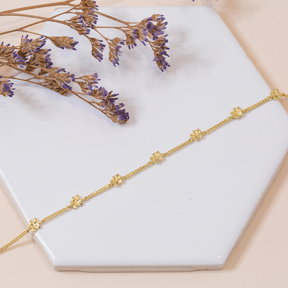 Our Hannah 7 almond blossoms Christian bracelet in yellow gold plated displayed on a white tile with small purple dried flowers with a pale pink fabric background.